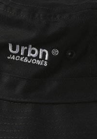 Black fabric bucket hat featuring embroidered "Urbn JACK&JONES" logo in white. Stitched accents and a textured finish throughout.