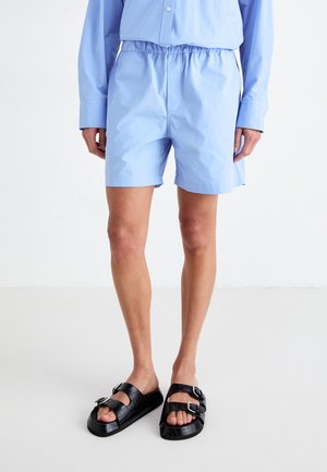 Lyseblå shorts laget av glatt stoff med elastisk midje. Kombinert med svarte sandaler med spenner som har en teksturert design.