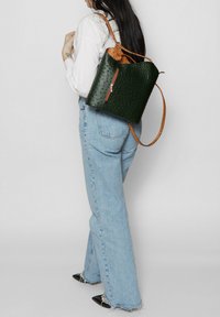 Sac à dos en cuir texturé vert avec des sangles en cuir marron, comportant une poche frontale zippée et une forme douce et arrondie.