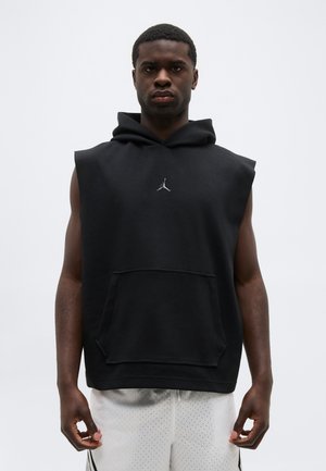 Giovane uomo con felpa nera senza maniche con logo Jordan e pantaloncini da basket bianchi traforati, in piedi davanti a uno sfondo semplice.