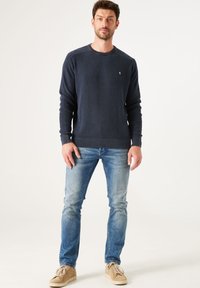 Marineblauer Strickpullover mit gerippter Textur, Rundhalsausschnitt und kleinem Logo. Kombiniert mit hellblauen Jeans und beigen Turnschuhen.