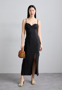 Robe noire ajustée avec un décolleté en cœur, une fente sur le côté et des bretelles spaghetti. Associée à des sandales à talons vertes et un sac à main tressé marron.