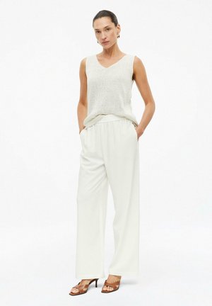 Femme debout, les mains dans les poches, portant un haut en maille beige sans manches, un pantalon large blanc et des sandales à talons marron sur fond blanc.