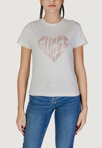 T-shirt bianco in cotone a maniche corte con una fantasia a forma di cuore realizzata in strass rosa e la parola "GUESS" in un carattere stilizzato.