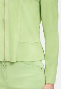 Jasnozielona kurtka o gładkiej teksturze, z zamkiem błyskawicznym i fasonem peplum, w połączeniu z dopasowanymi zielonymi spodniami z bocznymi kieszeniami.