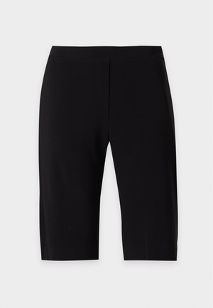Femme portant un chemisier en dentelle violette transparente à épaules dénudées, un short noir longueur genoux et des sandales noires à talons, debout avec une main sur la hanche.