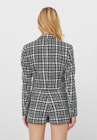 Blond kvinna som bär en svart- och vitrutigt kavaj och matchande shorts, stående med ryggen mot kameran.