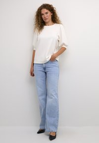 Wit, kortärmige top met een losse pasvorm, gecombineerd met lichtblauwe, wijde jeans en zwarte puntige schoenen op een effen achtergrond.