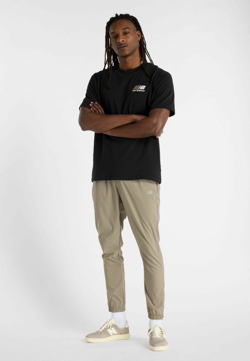 Camiseta negra con un logo, pantalones beige ajustados y zapatillas ligeras. El conjunto presenta un ajuste relajado y puños elásticos en los pantalones.