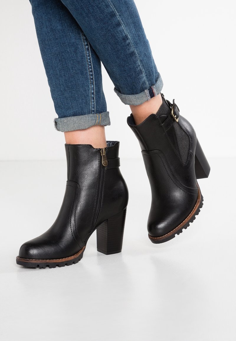 Bottines en simili cuir noir avec un talon épais, fermeture éclair sur le côté et couture ton sur ton. Présente une sangle avec un détail en métal doré.