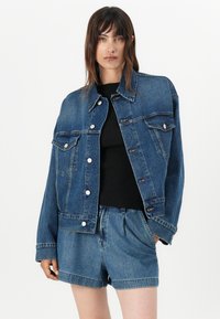 Denim jakna u tamnoplavoj boji, sa klasičnim dizajnom, dugmadi, džepovima i opuštenim krojem. Nosena preko crne majice i kratkih hlača.