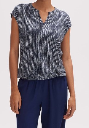 Vrouw die marineblauwe broek draagt en een marineblauw shirt met korte mouwen, voorzien van een wit abstract stippenpatroon en een kleine V-hals opening.