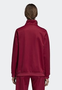 Rote Sportjacke mit hohem Kragen, glatter Textur und geripptem Saum. Die Ärmel sind lang und haben eine lockere Passform im Rücken.