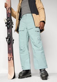 Pantaloni de zăpadă de un albastru deschis, cu buzunare cargo, manșete strânse și branding Columbia, asortați cu cizme de schi negre și o placă de snowboard.