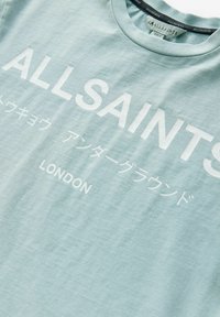 Vaaleansininen puuvillapaita, jossa on suuri valkoinen teksti "ALLSAINTS" ja "LONDON" sekä lisäkirjaimia japaniksi, pyöreä kaula-aukko.
