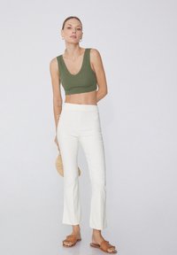 Olivově zelený krátký top bez rukávů, bílé rozšířené kalhoty a hnědé sandály s třásněmi. Modelka drží tkanou slámovou tašku, minimalistické pozadí.