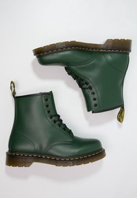 Zielone gumowe botki za kostkę z czarnymi sznurowadłami, żółtym przeszyciem i pętelką do łatwego zakładania. Gładka tekstura, solidna podeszwa i zaokrąglony czubek.