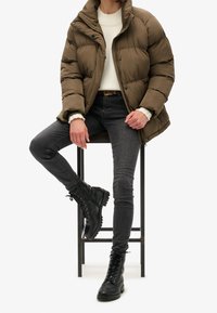 Bruine pufferjas met een hoge kraag, gedragen over een witte trui. Gecombineerd met donkere jeans en zwarte vetercombatlaarzen.