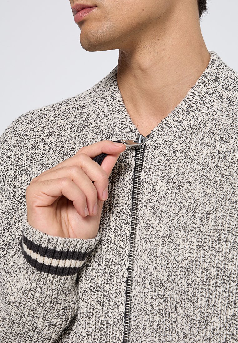 Pull en tricot gris et blanc avec fermeture éclair. Présente des poignets côtelés à rayures noires et blanches. Gros plan montrant une main tenant le tirette de la fermeture éclair.