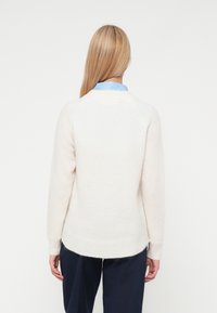 Ξανθιά γυναίκα που φοράει ένα λευκό πουλόβερ με μακριά μανίκια πάνω από ένα μπλε πουκάμισο, στέκεται με την πλάτη προς την κάμερα σε έναν απλό φόντο.