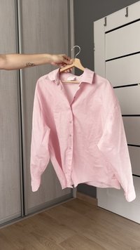 Pink striped button-up shirt with a classic collar, long sleeves, and a relaxed fit, hanging on a wooden hanger against a neutral backdrop.