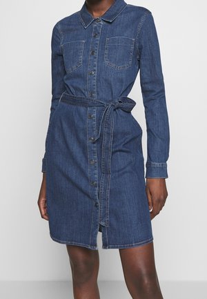 Kobieta ubrana w dżinsową sukienkę do kolan z długim rękawem, z przodem na guziki, dwoma kieszeniami na piersi i paskiem przewiązanym w talii.