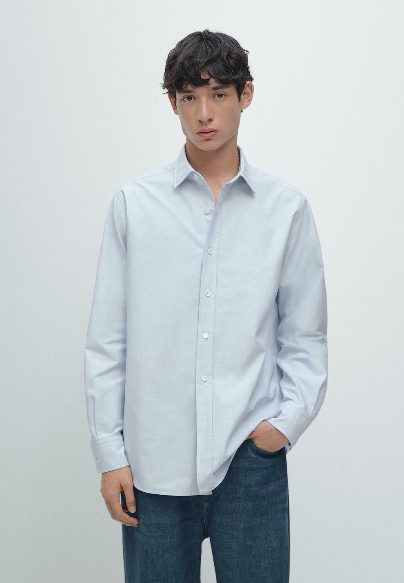 Jasnoniebieska koszula z kołnierzykiem, z długimi rękawami i luźnym krojem, zestawiona z ciemnoniebieskimi dżinsami. Gładka, wypolerowana faktura materiału.