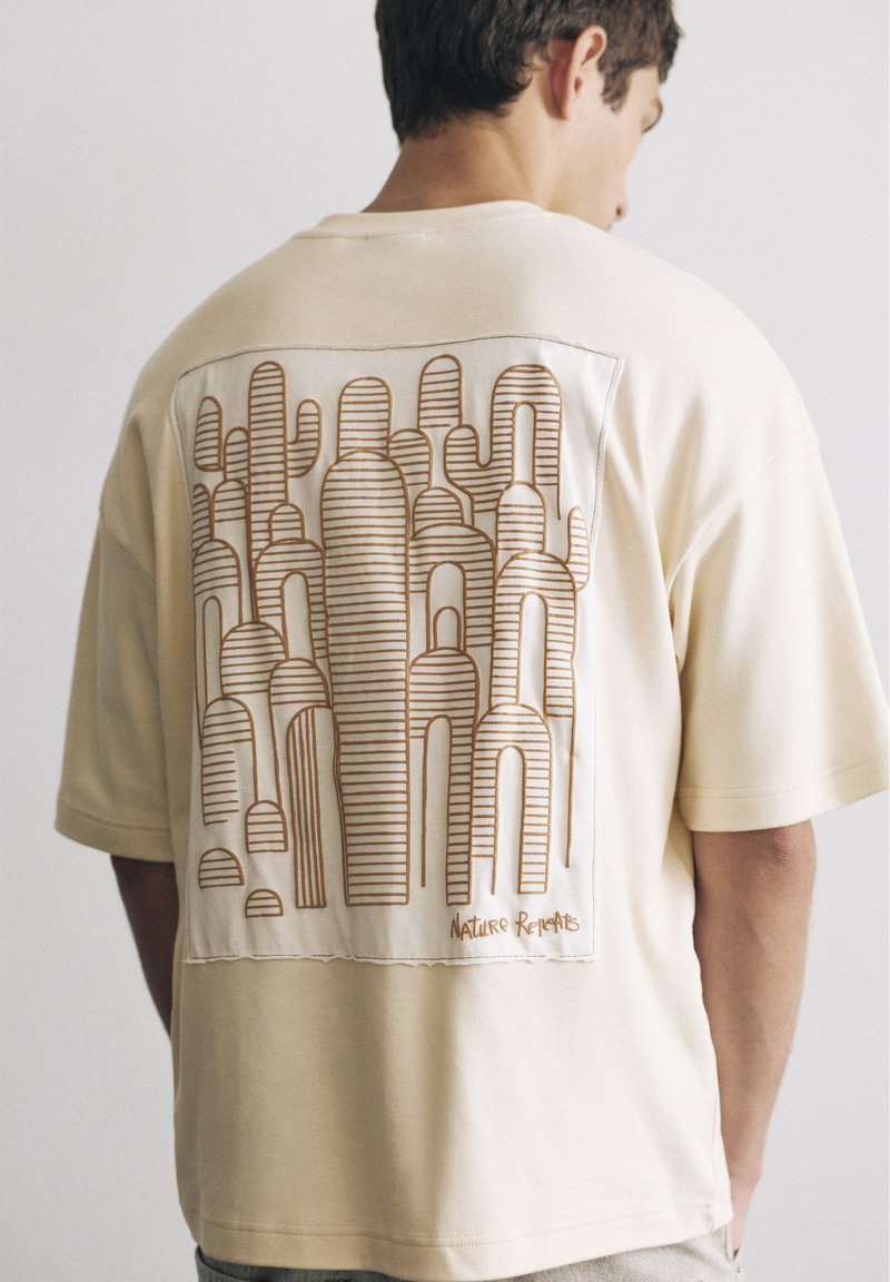 Homme portant un t-shirt beige avec un dessin au trait marron de cactus stylisés et le texte "Nature Relents" dans le dos.