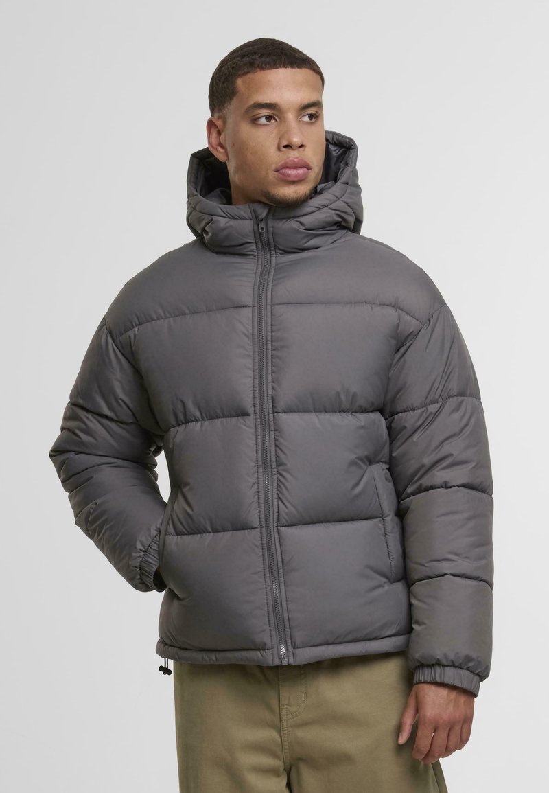 Grå pufferjakke med hette, med glidelås, elastiske mansjetter og et quiltet design. Mykt, lett materiale med en glatt tekstur.