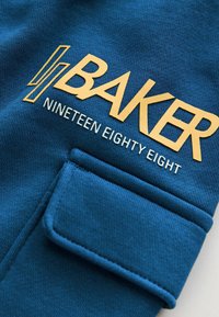 Blauwe stof met een getextureerd breipatroon; bevat "BAKER" en "NINETEEN EIGHTY EIGHT" in gouden letters; inclusief een klein pocketdetail.