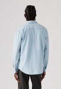 Man met vlecht haar in een lichtblauw denim overhemd met lange mouwen en donkere broek, staande met de rug naar ons toe tegen een effen achtergrond.