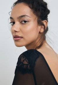 Artimas plano moters su tamsiais, bangomis plaukais, vilkinčios permatomą juodą marškinį su nėrinių akcentais, pabrėžiančiu jos odos atspalvį ir natūralią makiažą.