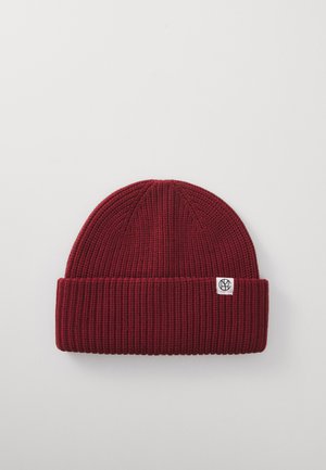 Dark red ribbed knit beanie with folded cuff and small white tag featuring a black circular logo, displayed on a plain light background.