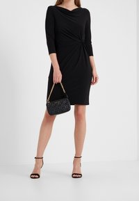 Kobieta w czarnej sukience do kolan z rękawami trzyczwartowymi, trzymająca małą czarną pikowaną torebkę na złotym łańcuszku.