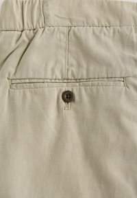 Tkanina khaki z prostokątną tylną kieszenią z brązowym zapięciem na guzik i wyraźnymi detalami przeszyć. Miękka tekstura, minimalistyczny design.