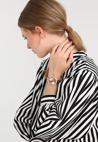 Różowa złota zegarek na rękę z okrągłą tarczą, minimalistycznym designem, gładką cyferblatą, połączonym paskiem, noszony na ręce w luźnej, prążkowanej czarno-białej tkaninie.