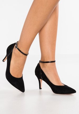 Vrouwenbenen met zwarte suède pumps met puntige neus, dunne enkellintjes en slanke middenhoge hakken op een witte vloer.