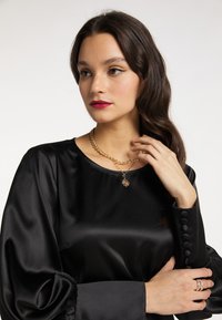 Sort silkebluse med lange, puffede ærmer og knapdetaljer. Guld kæderekæde med vedhæng. Modellens hånd hviler på hendes krave.