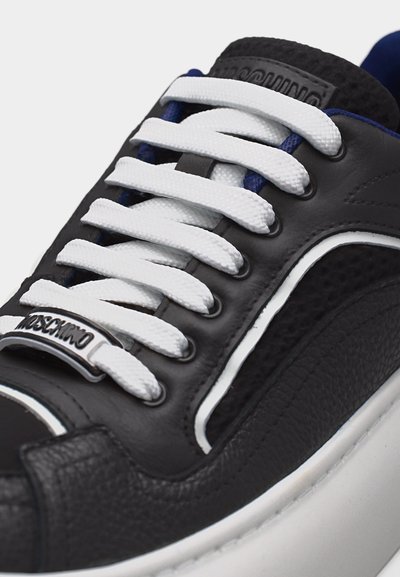 Juodas sportinis batelis su storomis baltomis raištelėmis, tekstūruotais odos skyriais, baltu padu ir metaline plokštele ant priekinių raištelių srities, išgraviruotu užrašu „Moschino“.