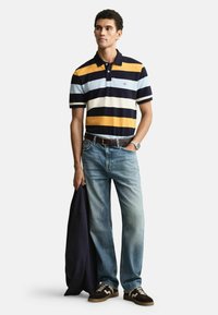 Koszulka polo w paski z krótkim rękawem w kolorze niebieskim, granatowym i pomarańczowym; luźne dżinsy; ciemna kurtka zarzucona na ramię; casualowe buty z teksturowanymi akcentami.