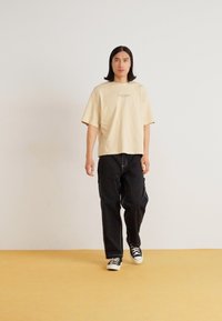 T-shirt beige surdimensionné avec un petit logo vert, associé à un pantalon cargo noir et des baskets noir et blanc, sur un fond uni.