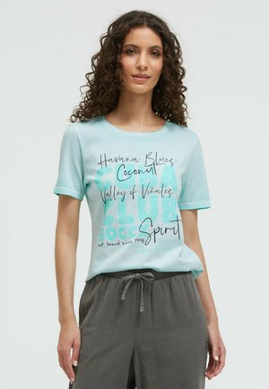 Junge Frau mit lockigem Haar, die ein hellblaues T-Shirt mit Grafik und graue Hose mit Kordelzug trägt, steht vor einem schlichten hellen Hintergrund.