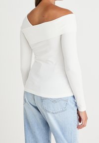 Žena v bílé dlouhé haleně s odkrytými rameny a světle modrých džínách s vysokým pasem, zobrazená zezadu.