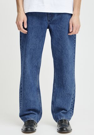 Mand iført løstsiddende blå cowboybukser, hvid skjorte og sorte lædersko, stående mod en ensfarvet lys baggrund.