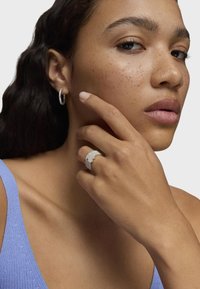 Anillo de plata con un diseño floral en relieve, llevado en la mano, combinado con un pendiente de aro de plata. El tono de piel parece uniforme con pecas.