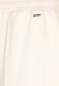 Bílé joggingové kalhoty s měkkou texturou, s elastickým pasem a malou stříbrnou označovací štítkou na boku.