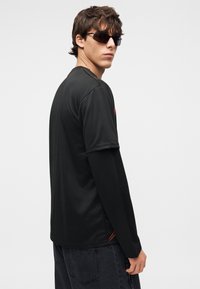 Νεαρός άνδρας φορώντας μαύρο πουκάμισο με μακριά μανίκια σε στρώσεις και μαύρα γυαλιά ηλίου, στέκεται πλάγια μπροστά σε απλό λευκό φόντο.