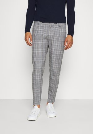 Mężczyzna w szarych, kraciastych, dopasowanych spodniach, białych trampkach i czarnej koszuli z długim rękawem stoi na tle białej ściany.