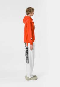 Sudadera con capucha naranja y cordones, combinada con pantalones blancos que presentan impresiones gráficas negras. El modelo lleva calzado ligero texturizado.