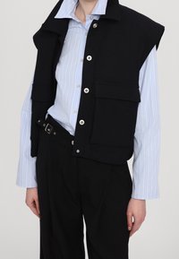 Zwarte mouwloze gilet met drukknopen en twee voorzakken, gedragen over een lichtblauw gestreept overhemd met knopen, gecombineerd met zwarte broek.